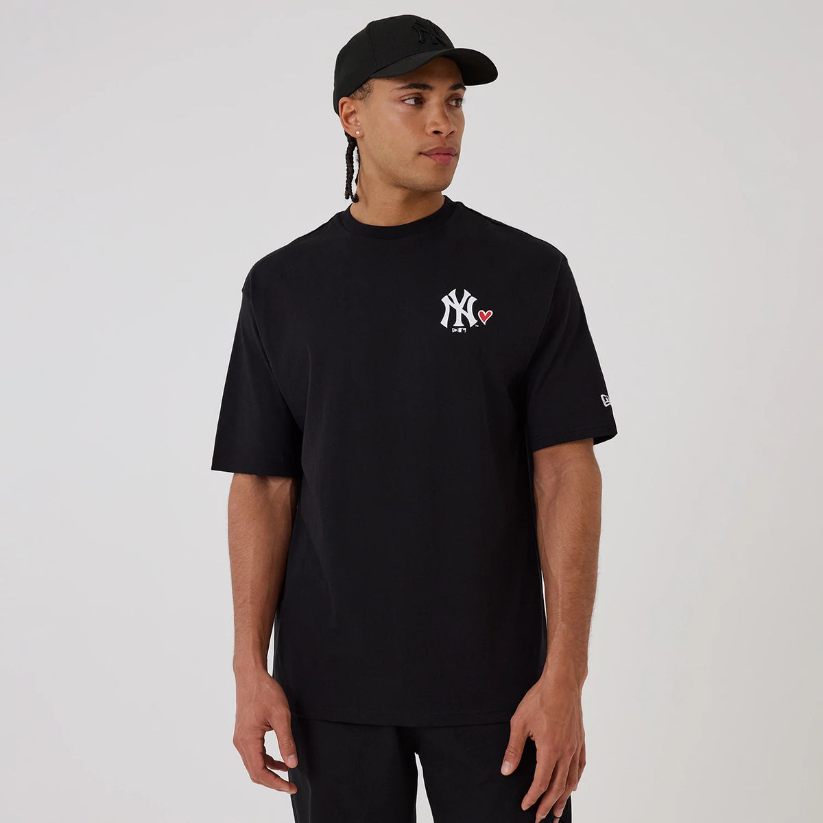 New Era New York Yankees MLB Heart Graphic Black T-Shirt, front view on a model with a matching black cap, showing the white embroidered NY logo with a small red heart on the left chest and a New Era logo on the left sleeve.
