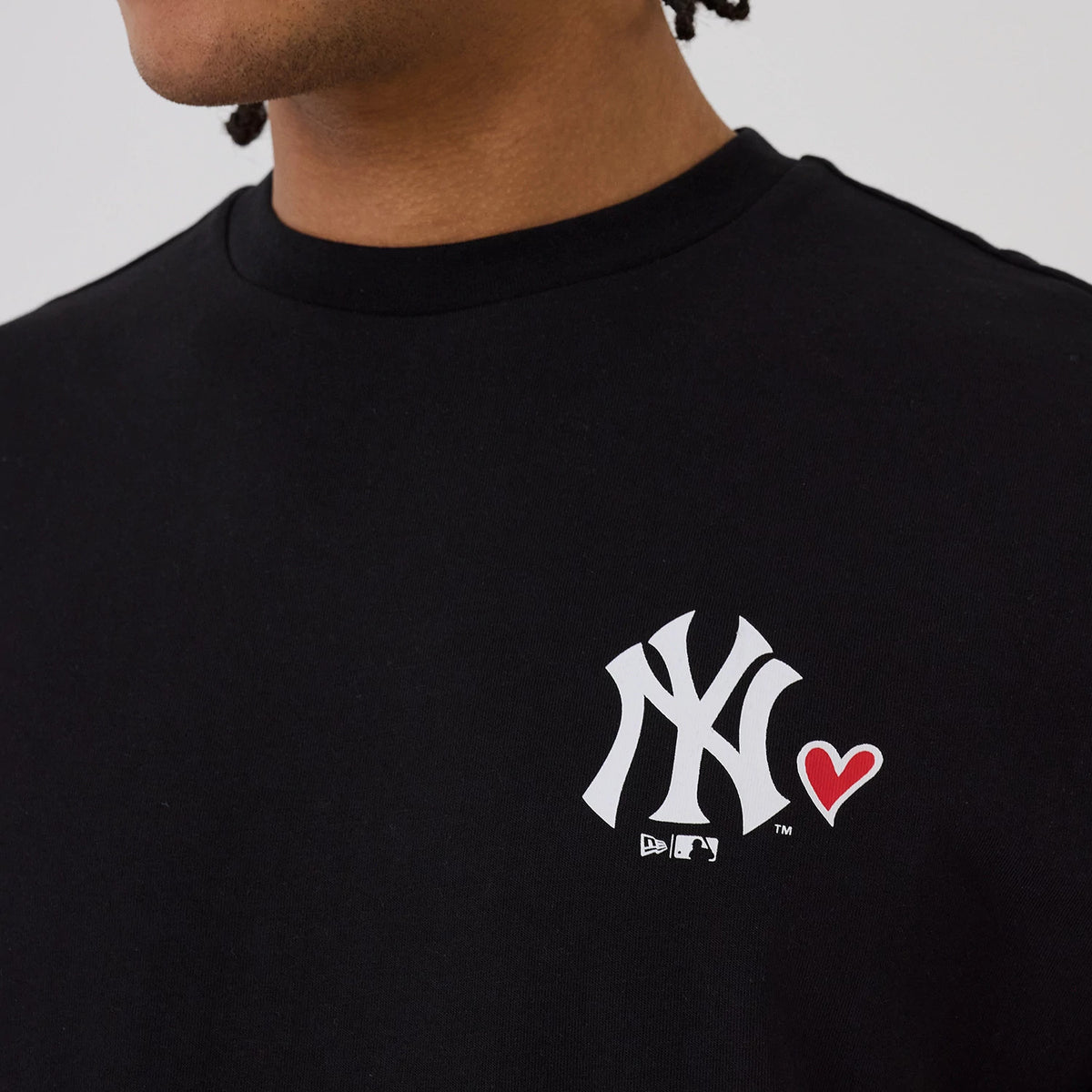 New Era New York Yankees MLB Heart Graphic Black T-Shirt, front view on-model showing a large white embroidered Yankees logo on the left chest with a small red heart graphic beside it, and a MLB logo underneath.