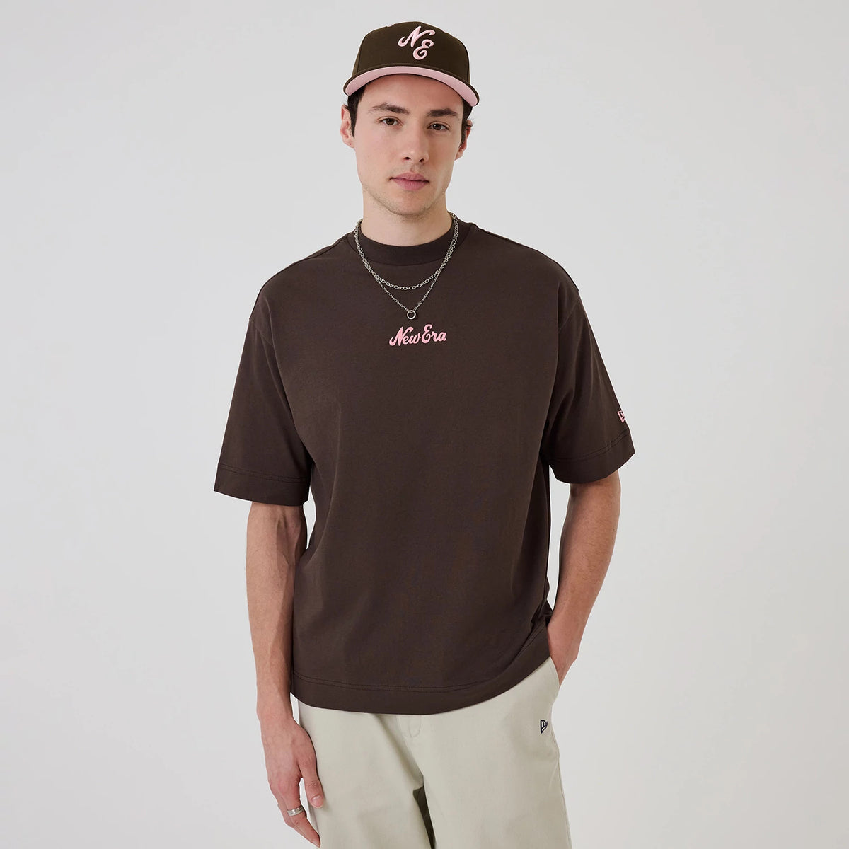 New Era Love Graphic Dark Brown T-Shirt, front view on-model showing a pink embroidered New Era logo centred on the chest, with short sleeves and an additional embroidered logo on the left sleeve, paired with a brown cap featuring pink accents.