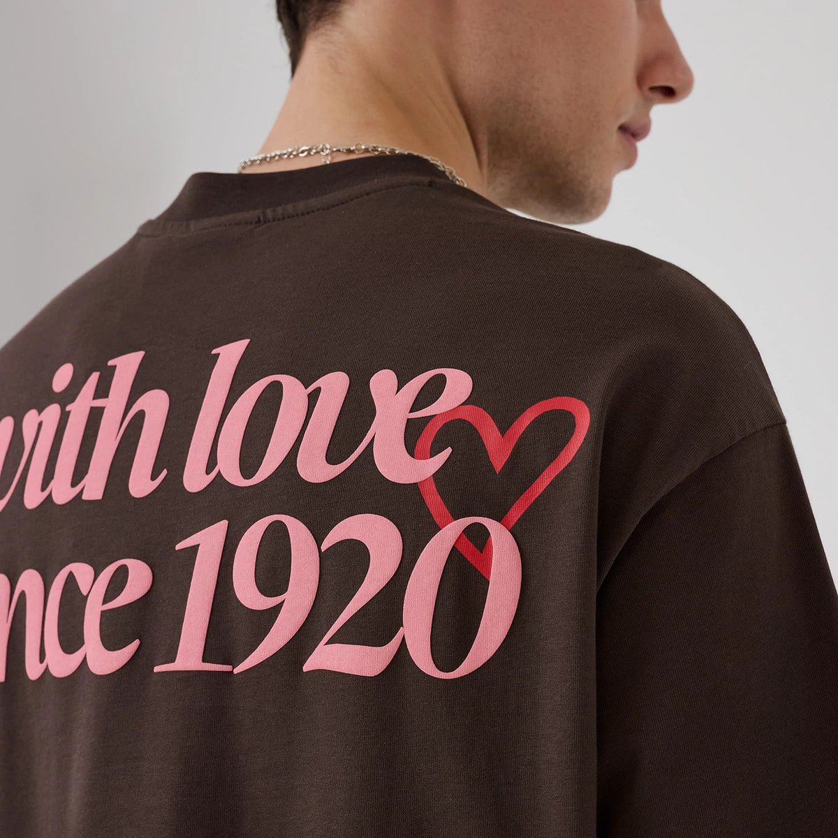 New Era Love Graphic Dark Brown T-Shirt, back view on a model showing pink lettering stating with love since 1920 across the upper back, featuring a red heart graphic intertwined with the text against the dark brown fabric.