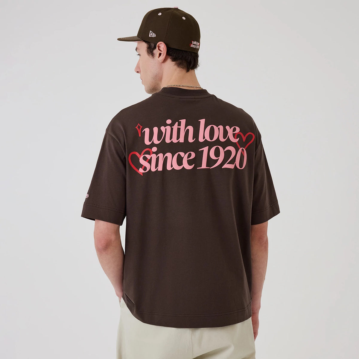 New Era Love Graphic Dark Brown T-Shirt, back view on a model showcasing pink printed with love since 1920 lettering with heart motifs, short sleeves and a New Era logo on the sleeve, paired with a dark cap and light trousers.