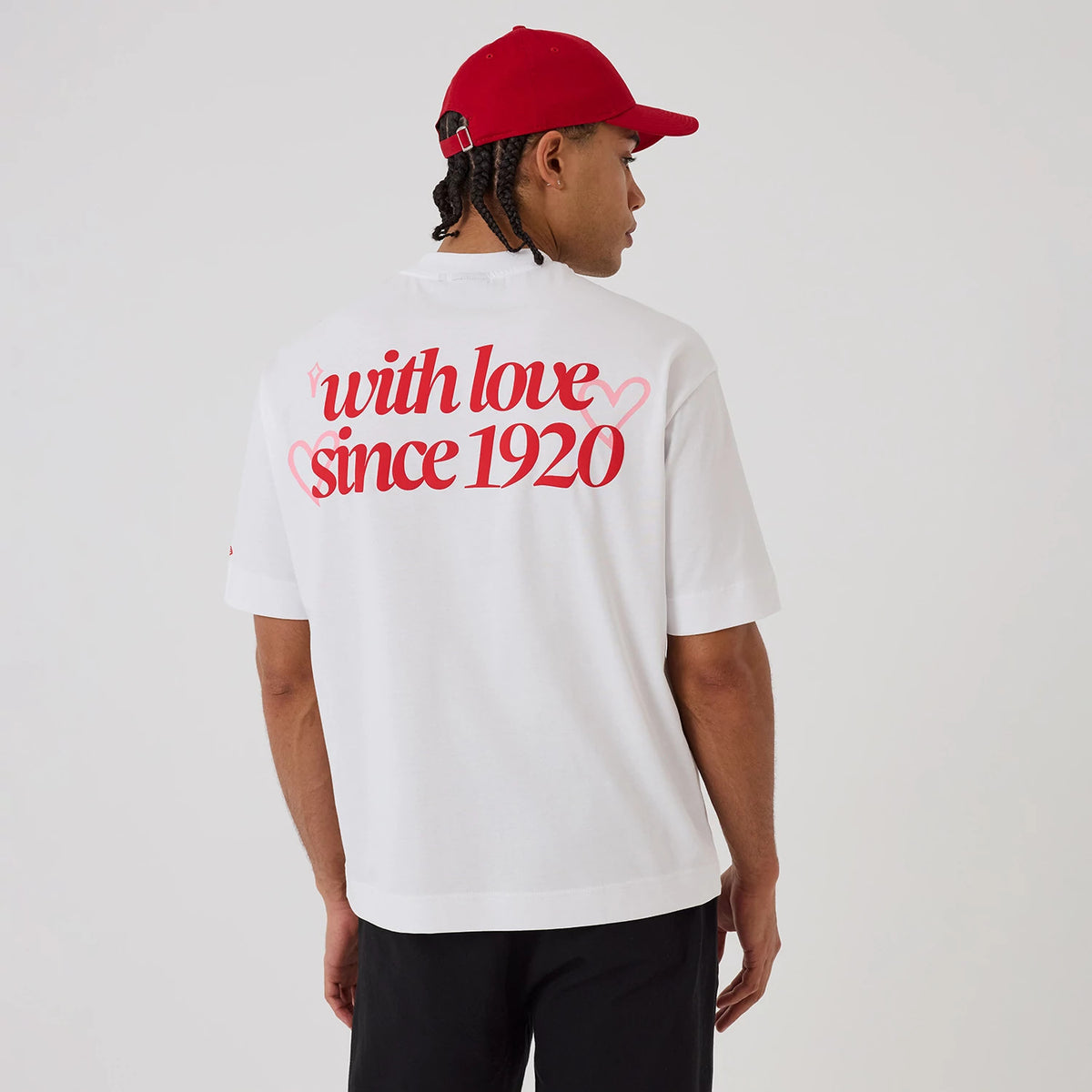 New Era Love Graphic White T-Shirt, back view on a model wearing a red cap, featuring the phrase with love since 1920 in red lettering across the upper back, with a pink heart graphic intertwined within the text.