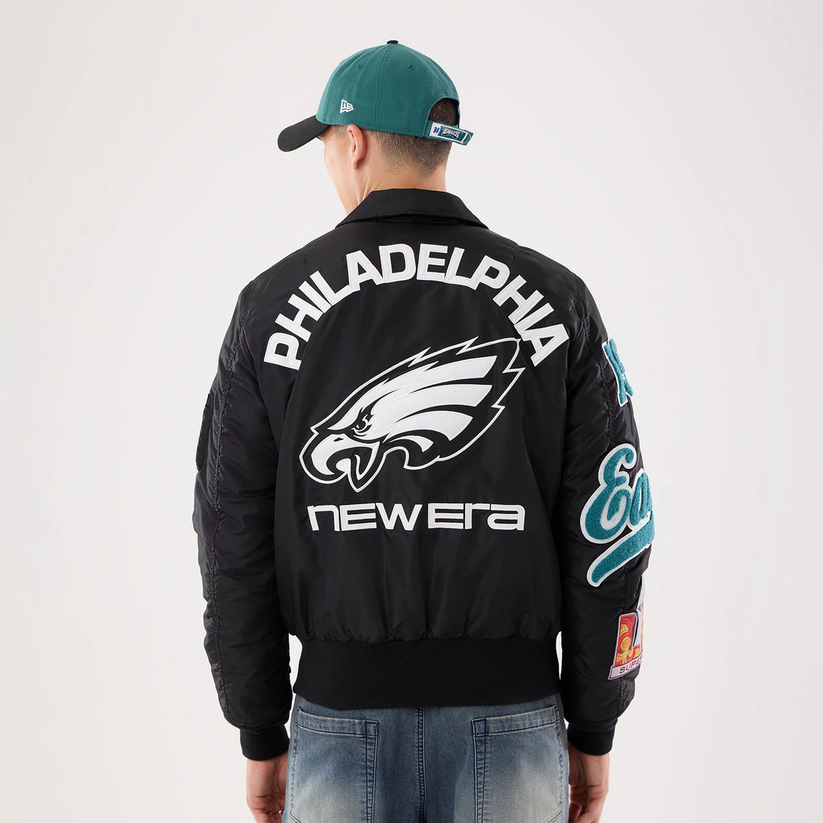 New Era Philadelphia Eagles NFL X Alpha Black Jacket, back view on a model wearing a green cap, showing a large Eagles logo in white with Philadelphia lettering above, and New Era branding below, with felt Eagles patch on the sleeve.