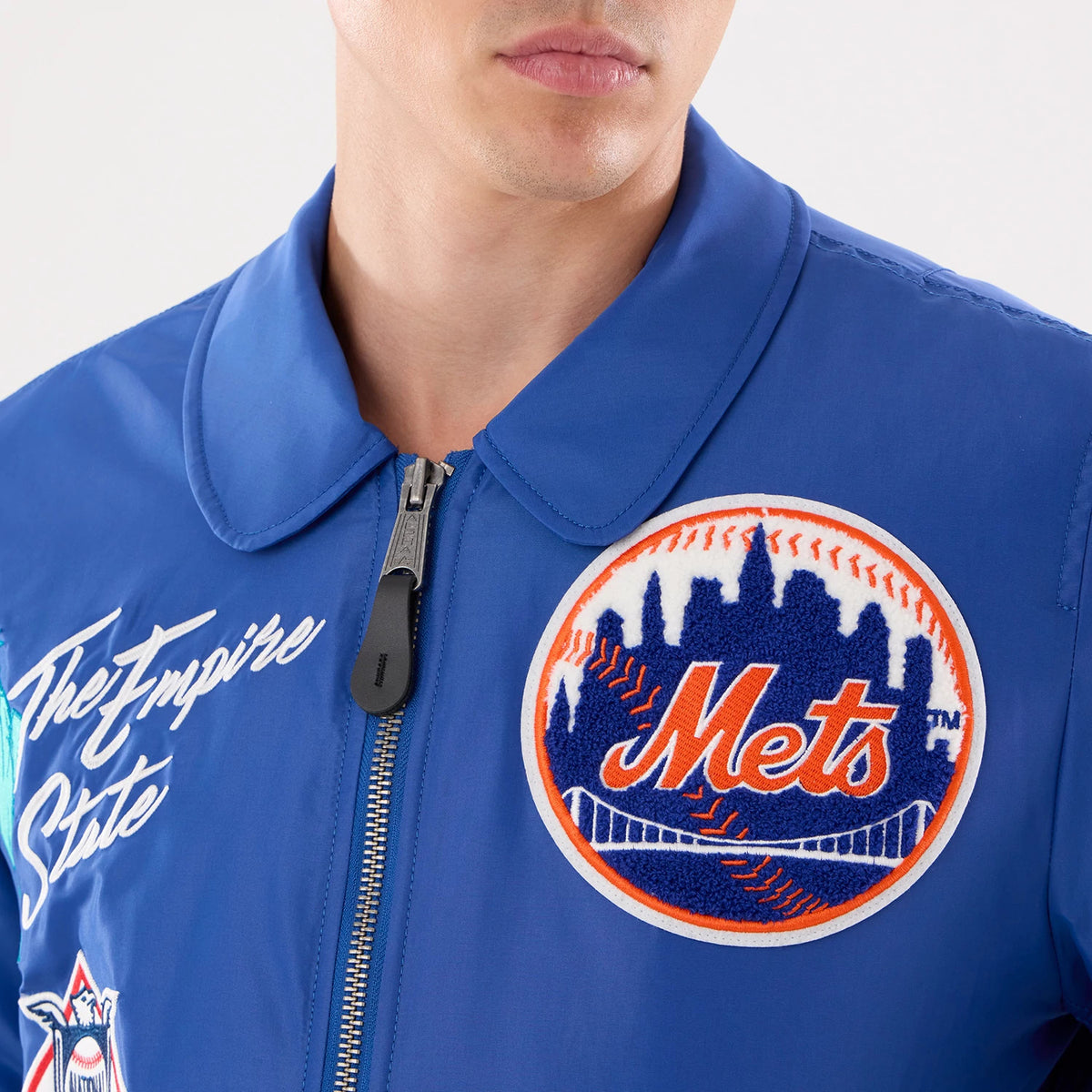 New Era New York Mets MLB X Alpha Blue Jacket, front view showing a large embroidered Mets logo on the left chest, a script 'The Empire State' embroidered on the right, and a central zip closure with a round collar on a blue jacket.