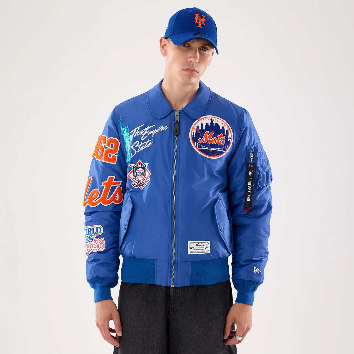 New Era New York Mets MLB X Alpha Blue Jacket, front view on a model, featuring embroidered Mets patches on the chest and sleeve, orange Mets lettering on the right arm, and a prominent zip pocket on the left sleeve with a branded tag.