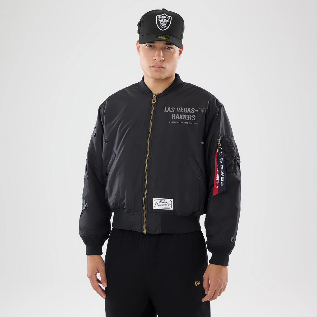 New Era Las Vegas Raiders NFL X Alpha Black Bomber Jacket & Tactical Vest, front view on-model showing the black bomber jacket with a zip closure, Las Vegas Raiders lettering on the chest, and a red tag on the sleeve, paired with a coordinating cap.