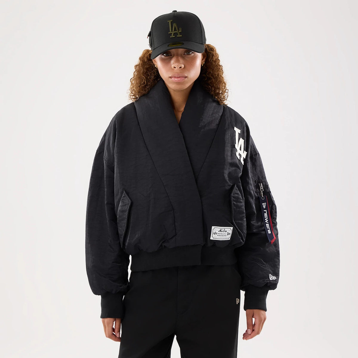 New Era LA Dodgers MLB X Alpha Black Jacket, front view on-model showing the oversized black design with an embroidered LA logo on the left chest, a patch above the ribbed hem, and a branded zip pocket on the sleeve with a red pull.
