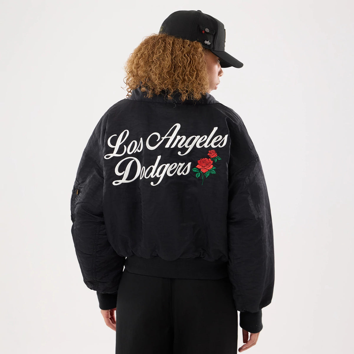 New Era LA Dodgers MLB X Alpha Black Jacket, back view showing large white embroidered Los Angeles Dodgers script across the shoulders, with red rose graphics below, model styled with a black cap and black trousers.