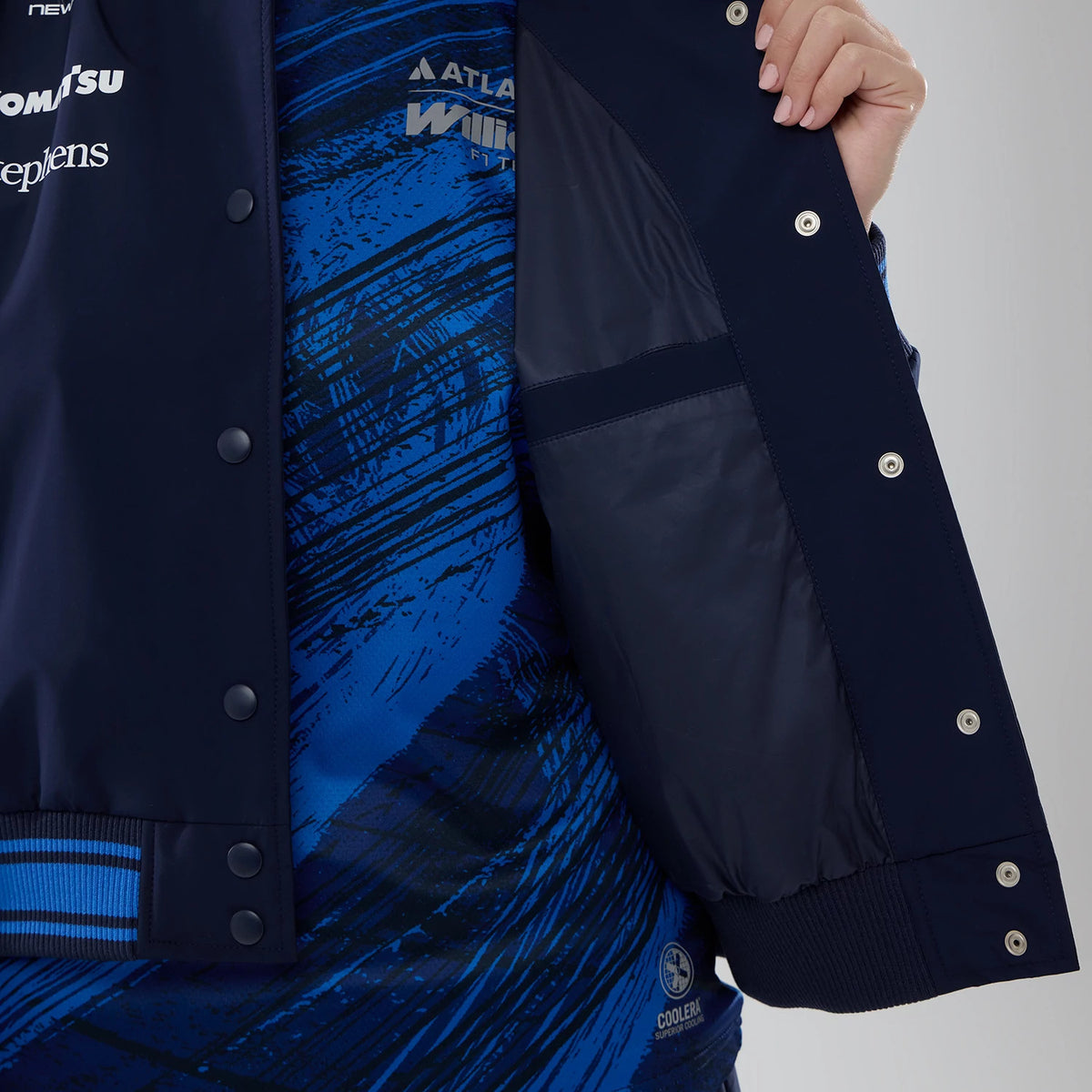 New Era Womens Williams Navy Varsity Jacket, inside view showing the jacket held open to reveal a navy lining with multiple snap buttons along the edge, worn over a blue patterned shirt with prominent graphics.
