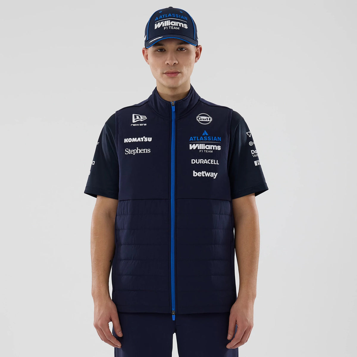 New Era Williams Navy Gilet, front view on a model with a matching Williams F1 Team cap, showcasing sponsor logos such as Komatsu, Stephens, and Duracell in white across the chest and shoulders, with a central blue zip closure.