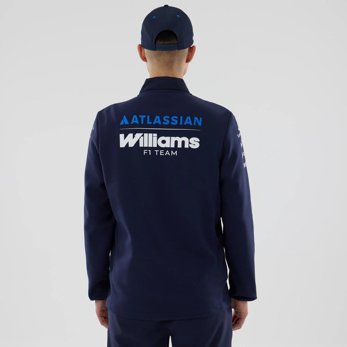 New Era Williams Navy Hybrid Jacket, back view on a model wearing a matching navy cap, featuring large white and blue printed Williams F1 Team and Atlassian logos on the upper back, with additional graphics on the sleeves.