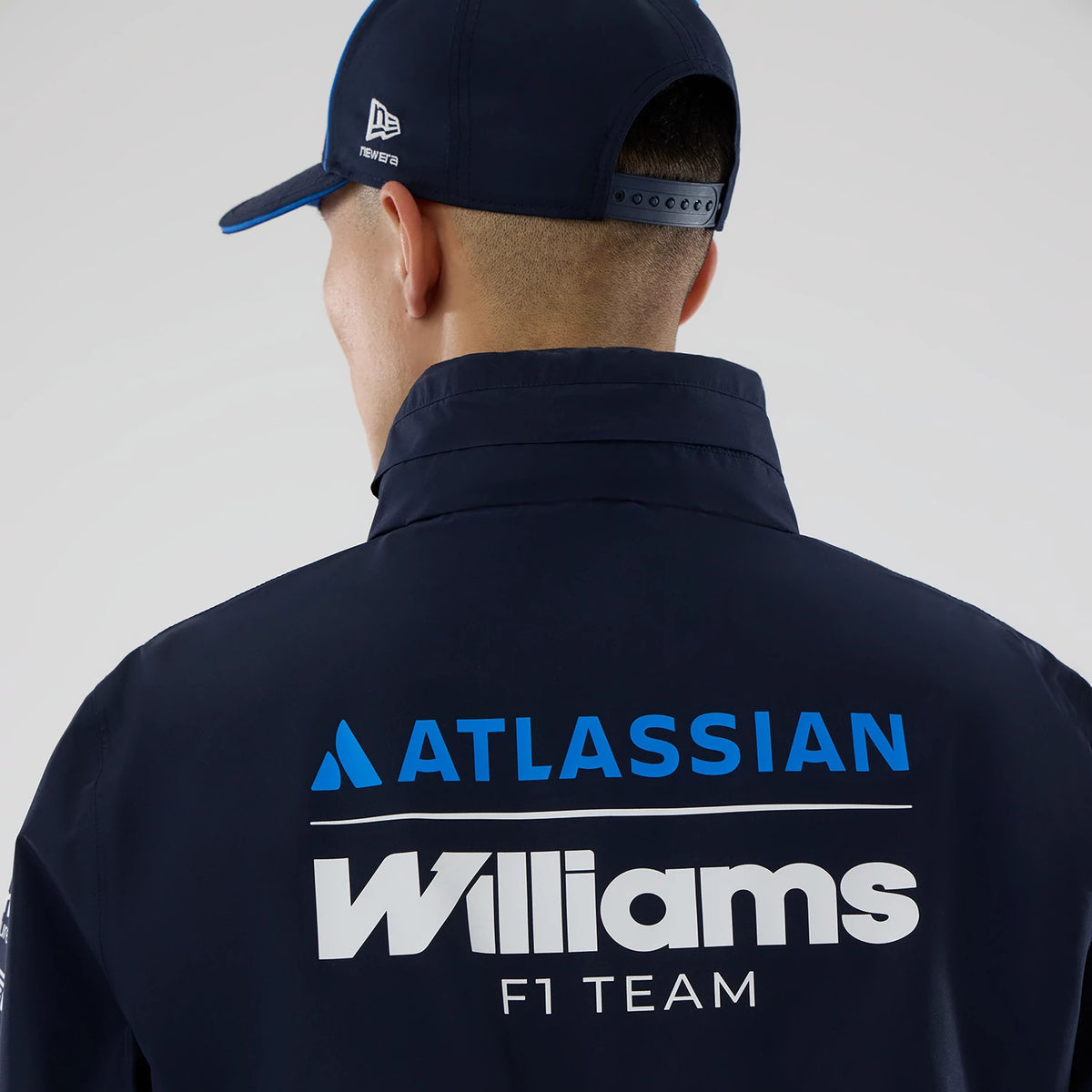 New Era Williams Navy Rain Jacket, back view on a model wearing a matching cap, featuring white and blue lettering of 'Atlassian' and 'Williams F1 Team' prominently printed across the upper back of the jacket in contrasting colours.