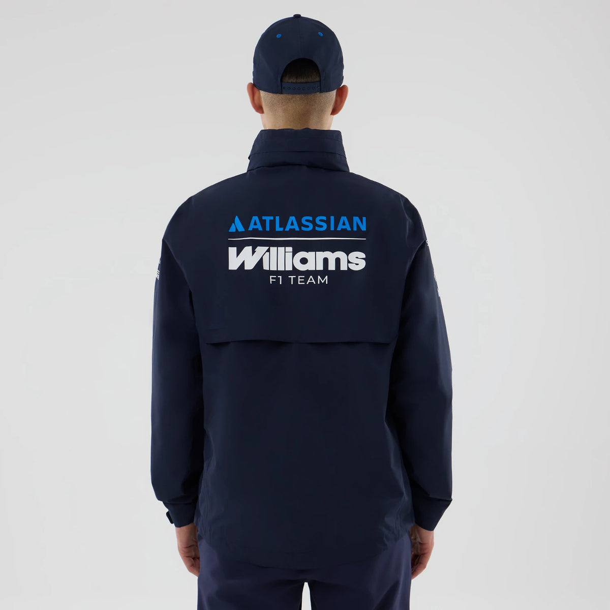 New Era Williams Navy Rain Jacket, back view showing prominent white Williams F1 Team lettering below a blue Atlassian logo, a flap at the back for styling on a model wearing a matching navy cap, enhancing the cohesive look of the ensemble.