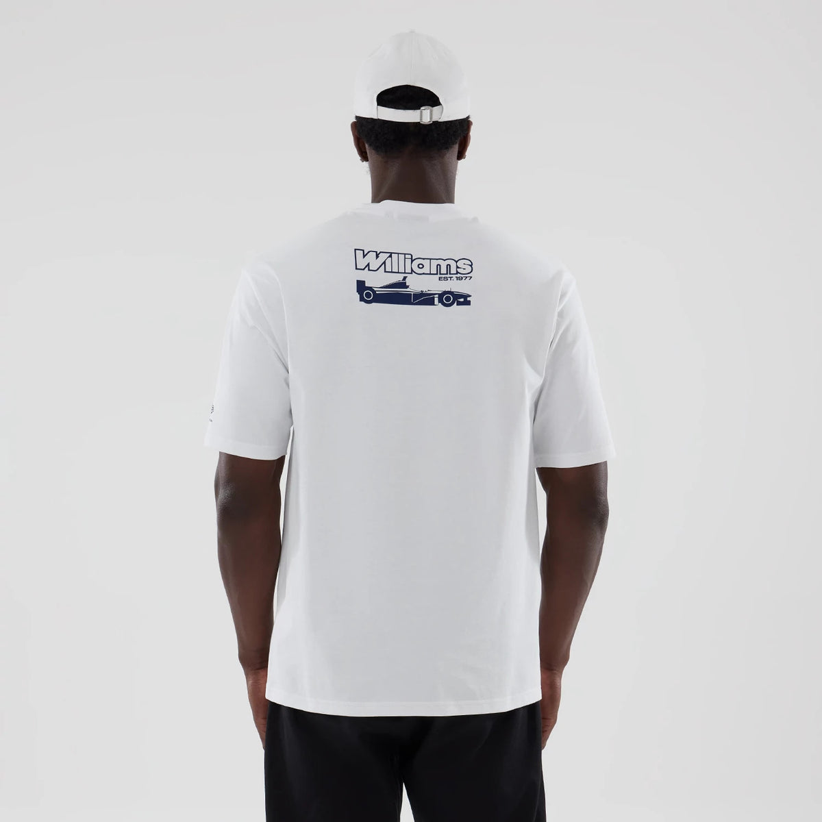 New Era Williams Japan 1996 White Oversized T-Shirt, back view on a model showcasing a blue printed graphic with Williams lettering and a formula car design centred on the upper back, styled with a white cap and black trousers.
