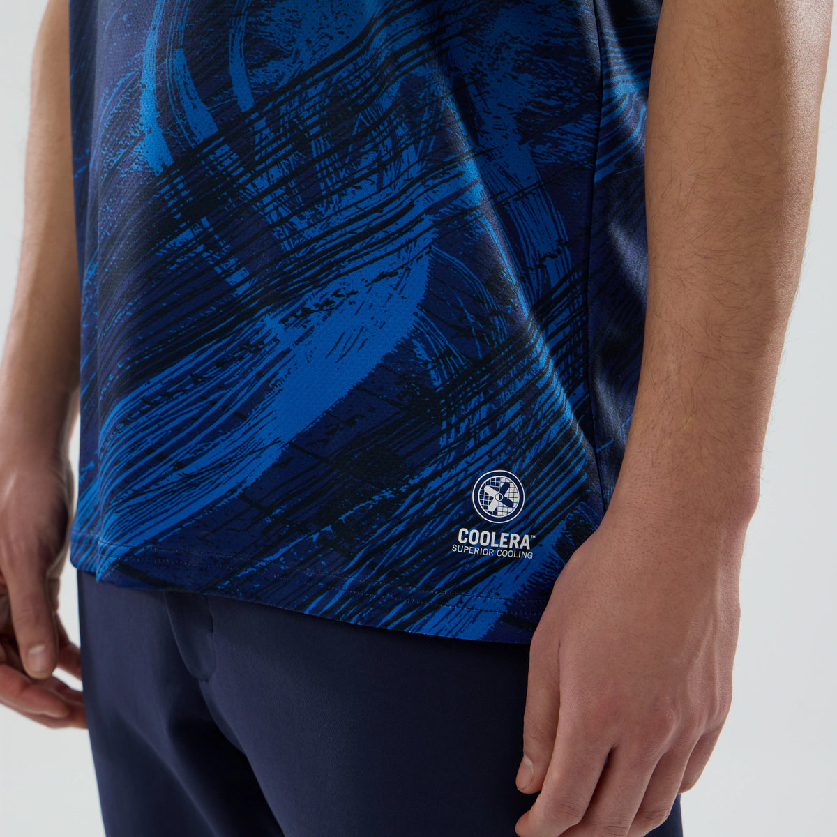 New Era Williams Carlos Sainz Navy Tech T-shirt, close-up view showing a blue and black brushstroke pattern across the fabric, a white printed Coolera logo on the lower side, and the hem partially above navy trousers.