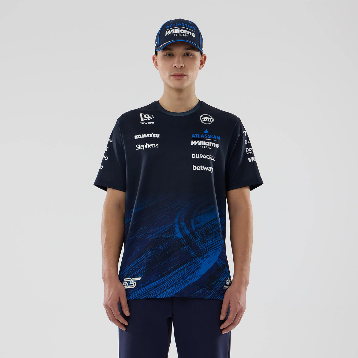 New Era Williams Carlos Sainz Navy Tech T-shirt, front view on a model paired with a blue cap, displaying multiple sponsor logos printed across the chest and sleeves, with a dynamic blue graphic pattern at the bottom.