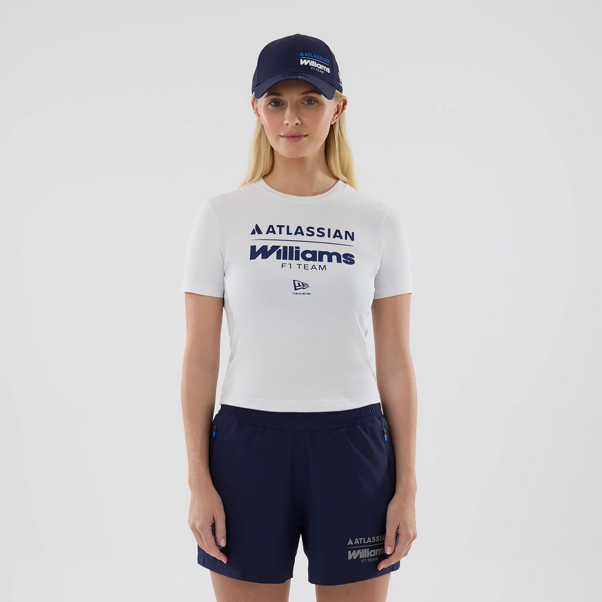 New Era Williams Womens White T-Shirt, front view on a model paired with a navy cap and navy shorts, featuring embossed Williams F1 Team lettering beneath an Atlassian logo, and an embroidered New Era flag logo centred below.