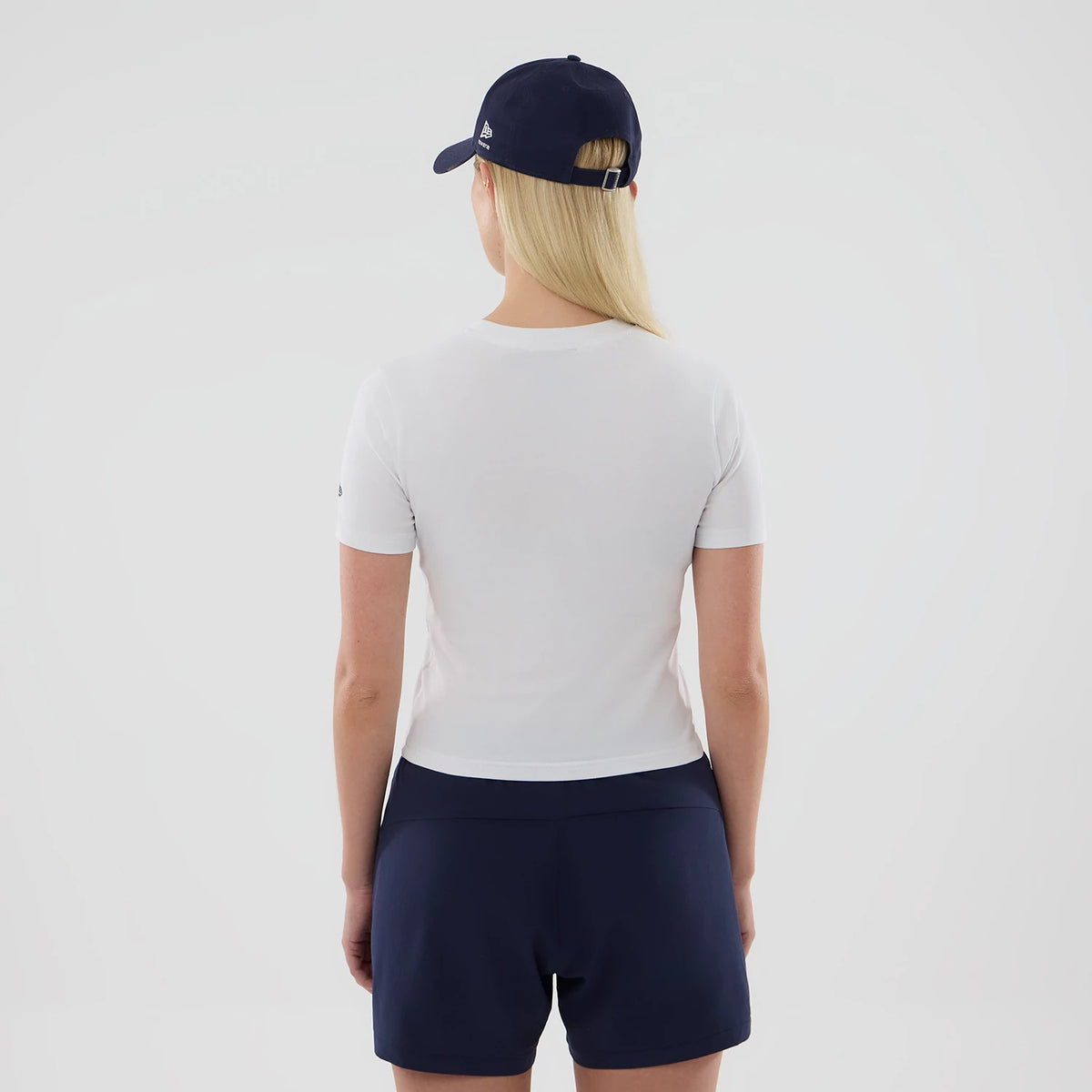 New Era Williams Womens White T-Shirt, back view on a model wearing a dark cap, showing the plain white short-sleeve design with a simple round neckline and a straight hem, complemented by navy shorts for a casual look.