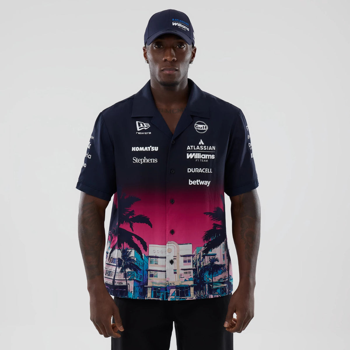 New Era Williams Miami Navy Short Sleeve Shirt, front view on a model with a navy blue cap, showcasing the navy and pink gradient design with palm tree graphics, multiple sponsor logos on both sides, and a button-up closure.