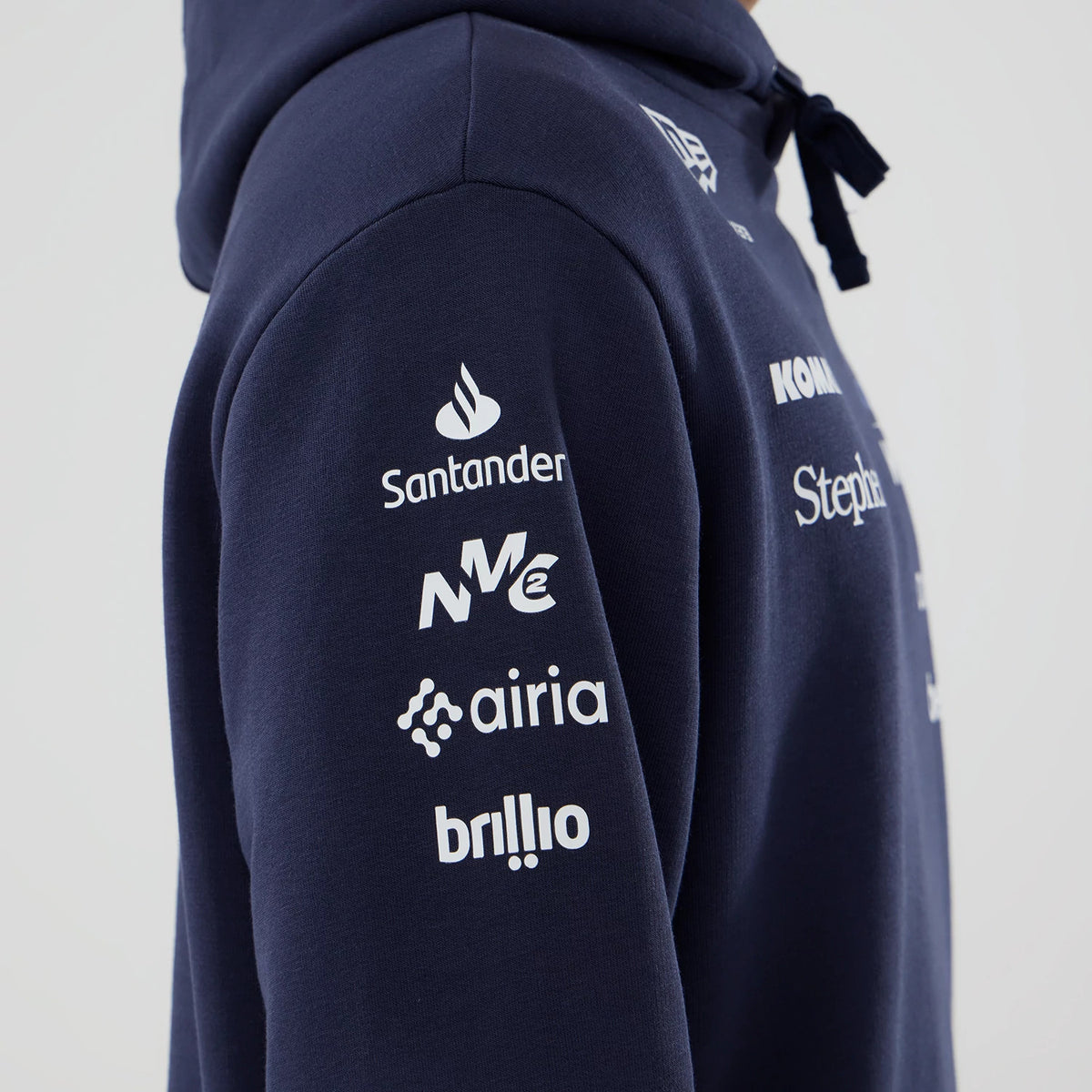 New Era Williams Replica Navy Pullover Hoodie, side view showing multiple white sponsor logos printed vertically down the sleeve, a New Era logo embroidered near the shoulder, and a drawn hood cord dangling at the front.