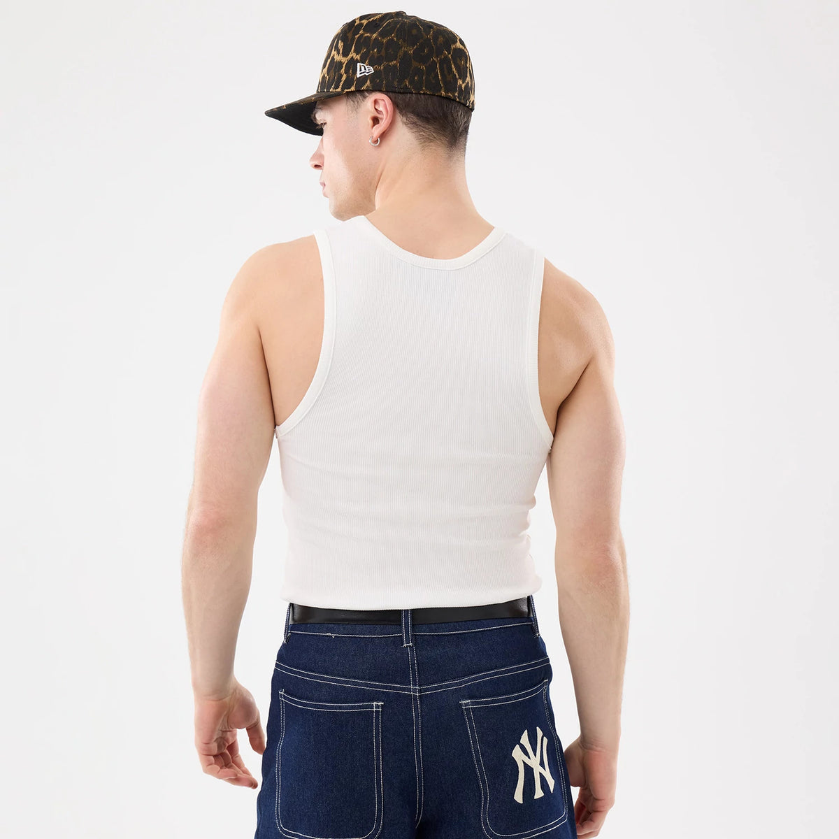 New Era New York Yankees MLB Rib White Vest, back view on a model wearing a leopard-print cap, showing the sleeveless design and ribbed texture of the white vest, paired with dark blue jeans featuring an embroidered NY logo on the back pocket.