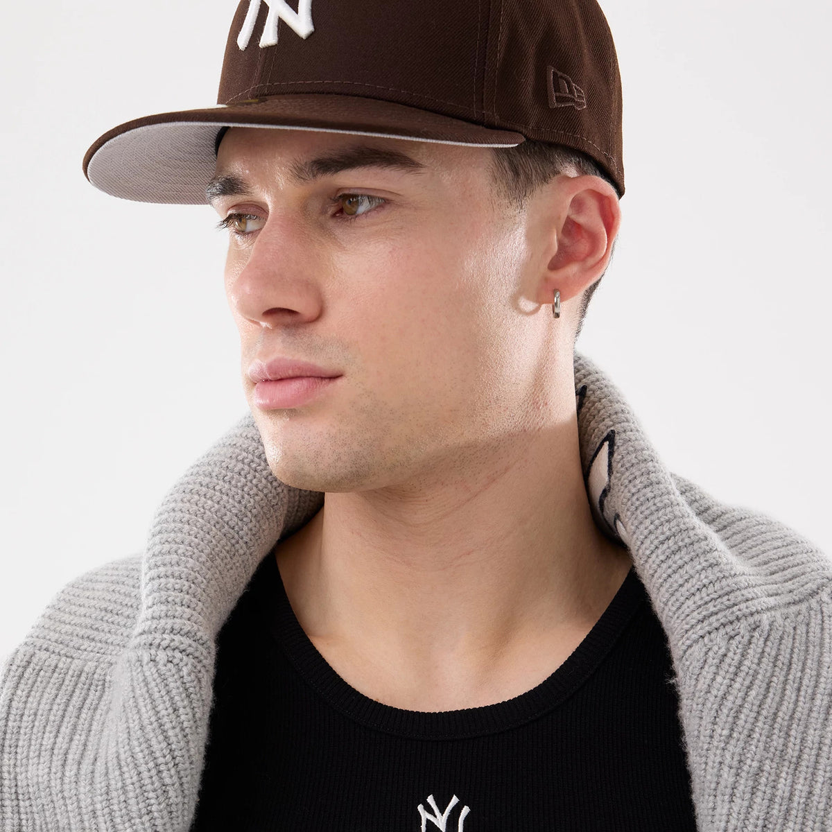 New Era New York Yankees MLB Rib Black Vest, on-model view with a close-up showing the black vest's upper design, featuring a white Yankees logo, alongside a brown cap worn by the model, and a grey knit collar draped over shoulders.