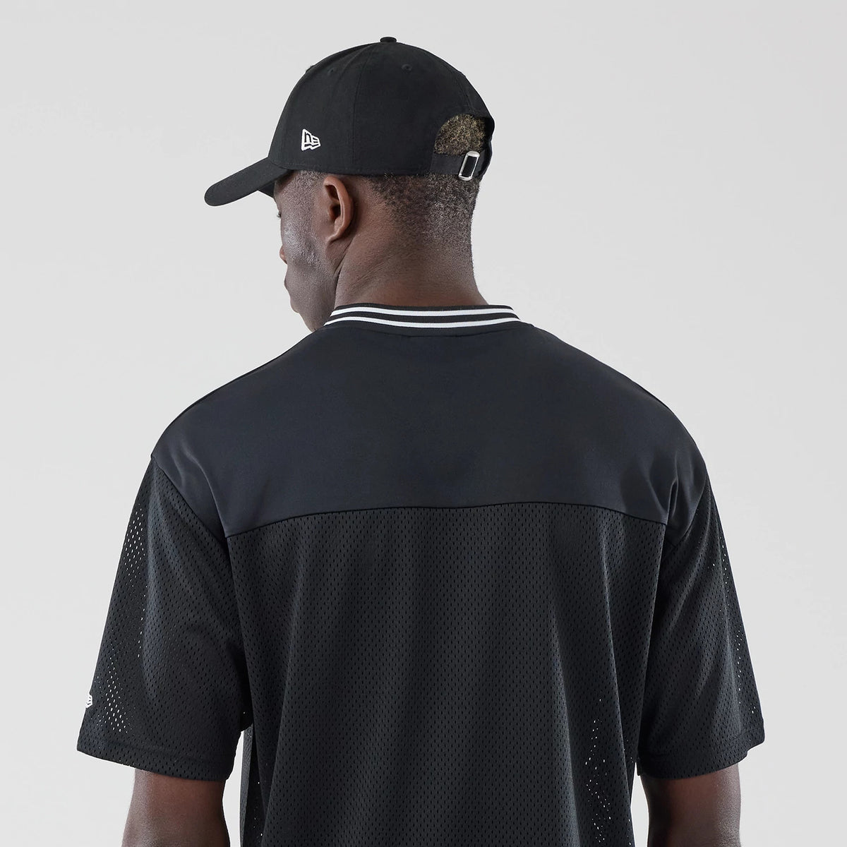 New Era Mesh Black Oversized T-Shirt, back view on-model showing the black fabric, contrasting white and black striped collar, and an embroidered New Era logo on the left sleeve, modelled with a black cap featuring a New Era logo.