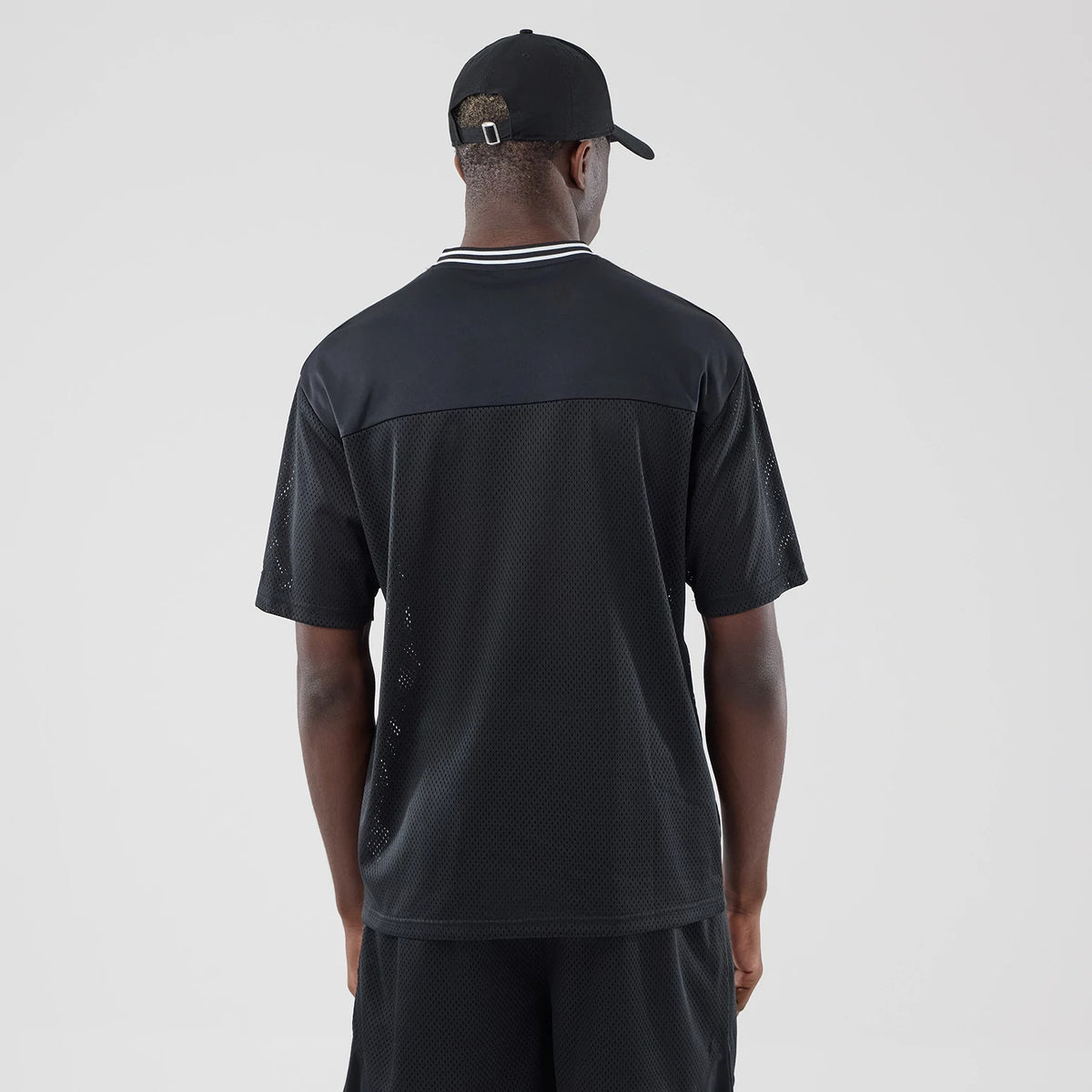 New Era Mesh Black Oversized T-Shirt, back view on a model showing the mesh construction of the shirt, a white-striped neckline, and short sleeves, paired with black shorts and a matching cap, creating a cohesive look.