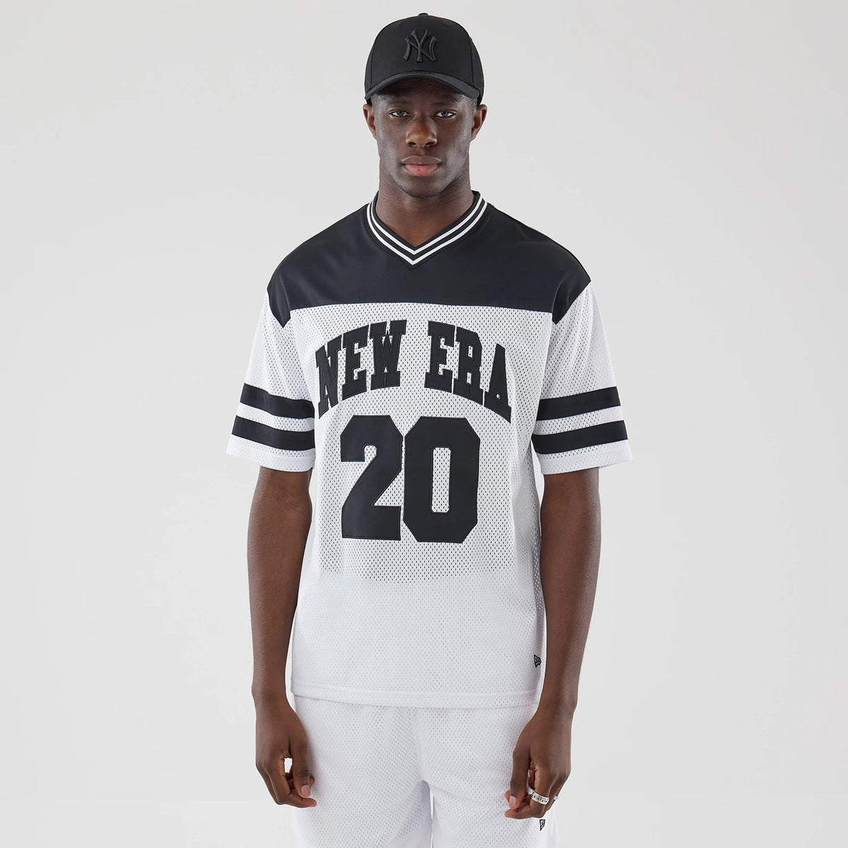 New Era Mesh White Oversized T-Shirt, front view on a model, featuring black 'NEW ERA' lettering across the chest, the number '20' printed below, and contrasting black stripes on the sleeves, styled with a simple black cap.