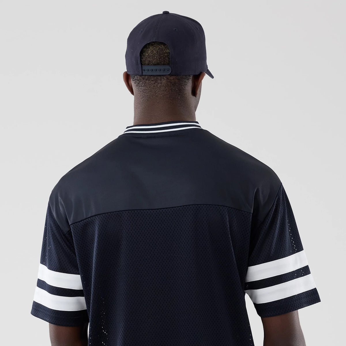 New Era Mesh Navy Oversized T-Shirt, back view on a model wearing a navy cap, showcasing the navy mesh fabric with contrasting white horizontal stripes on the sleeves and a double striped collar adding a sporty touch.