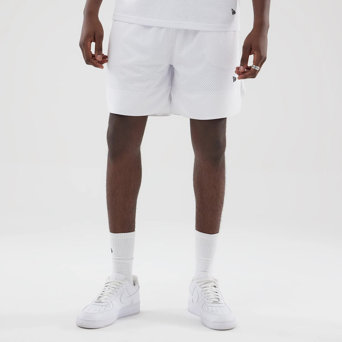 New Era Mesh White Oversized Shorts, front view on a model showing the knee-length design with a mesh texture, a embroidered New Era logo near the left hem, and paired with white trainers and socks for a coordinated look.