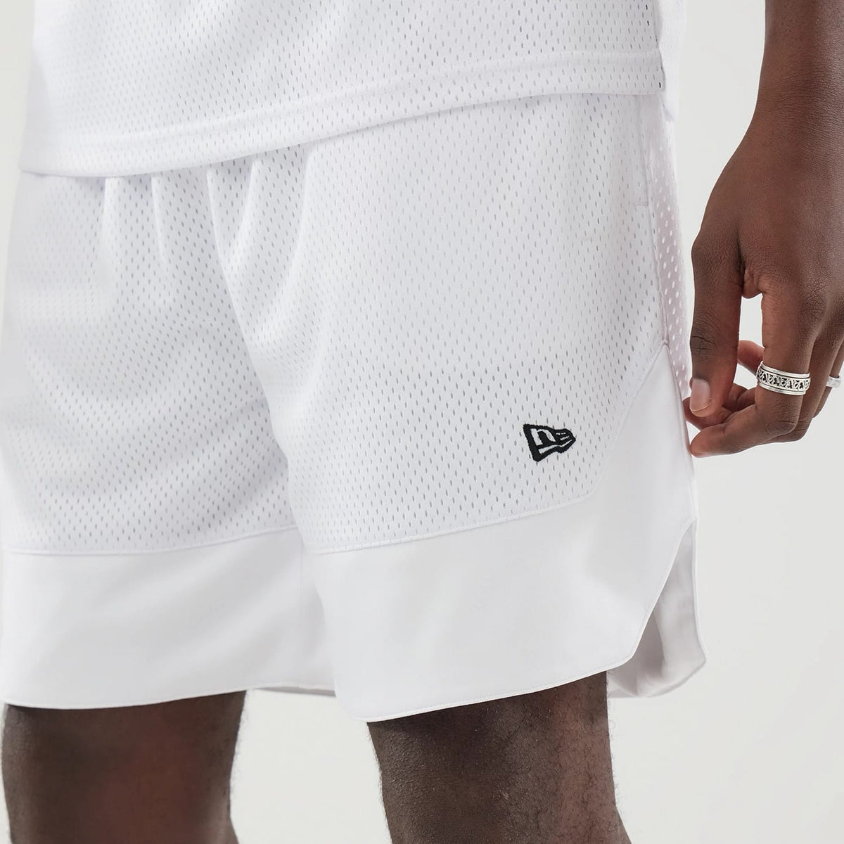 New Era Mesh White Oversized Shorts, front view showing the fine mesh texture with an embroidered black New Era logo on the lower left leg, a wide opaque hem at the bottom, and a relaxed fit on the model.