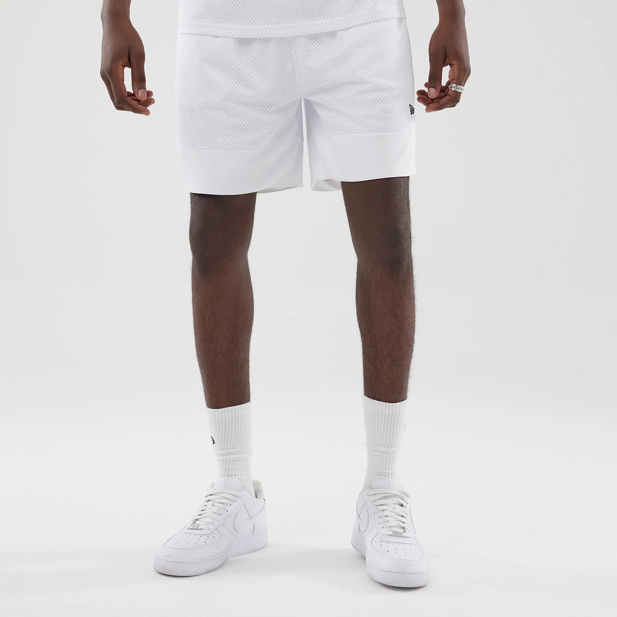 New Era Mesh White Oversized Shorts, front view on-model showing the white mesh fabric with a white hem, an embroidered New Era flag logo on the left leg near the seam, and styled with white socks and white trainers.