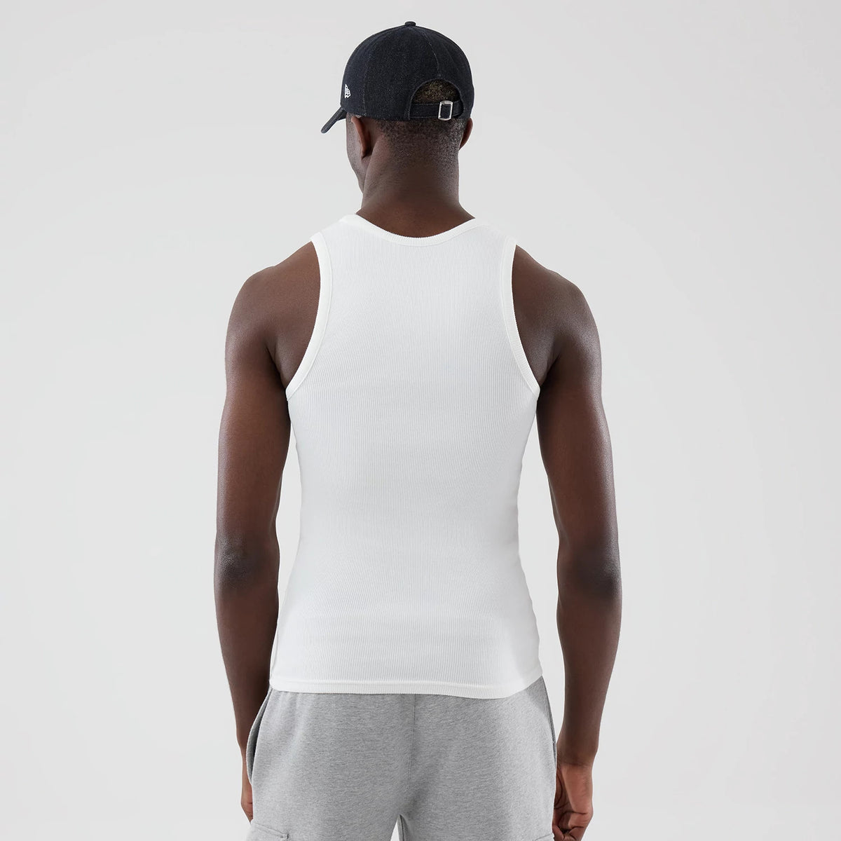 New Era Rib White Vest, back view on a model showing the ribbed texture of the white fabric, accentuated armholes, and a relaxed fit over grey shorts, complemented by a black cap with a New Era flag logo on the left side.