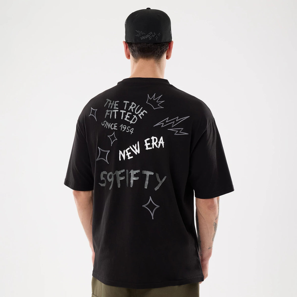 New Era 59FIFTY Day Graphic Black T-Shirt, back view on a model with a black cap, showcasing white graphics including THE TRUE FITTED, NEW ERA, and 59FIFTY, surrounded by stars and zigzag patterns across the upper back.