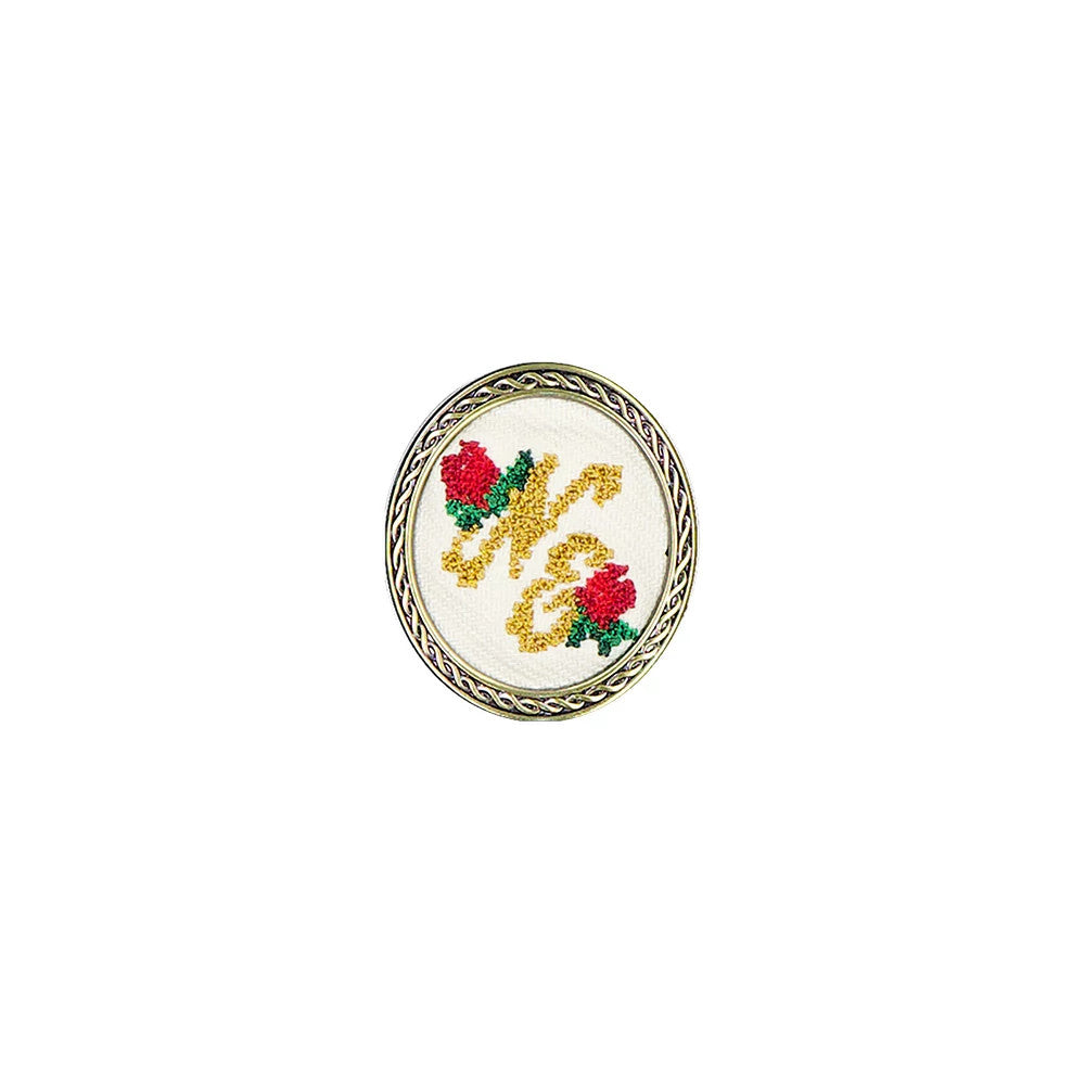 New Era 59FIFTY Day Needle Point Black Pin Badge, front view showing a circular design with gold needle point lettering and two red roses on a white backdrop, framed by a decorative silver border.