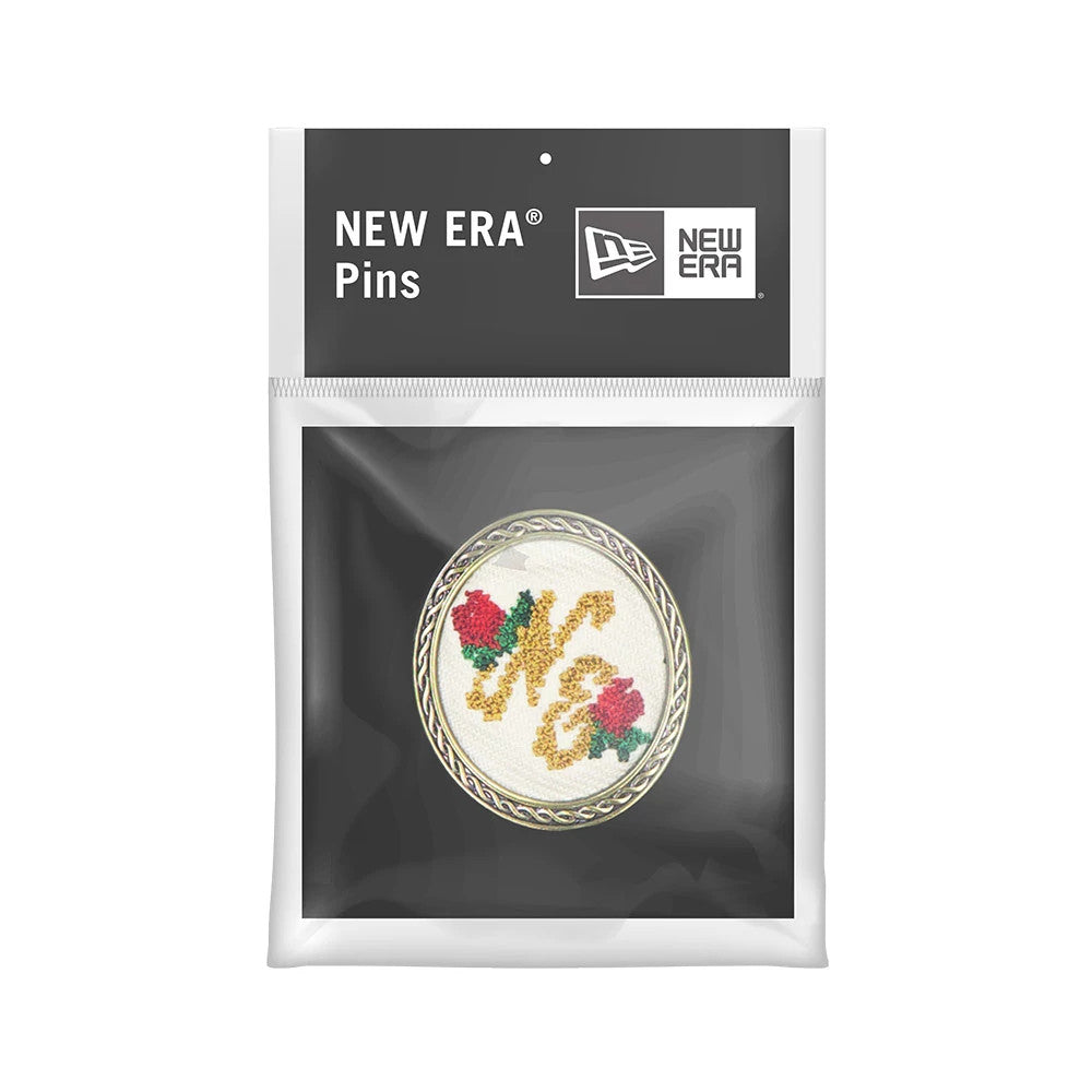 New Era 59FIFTY Day Needle Point Black Pin Badge, front view featuring an oval badge with intricate needle point design showing gold letters and red roses, encased in a twisted border, in a transparent package with New Era branding on top.