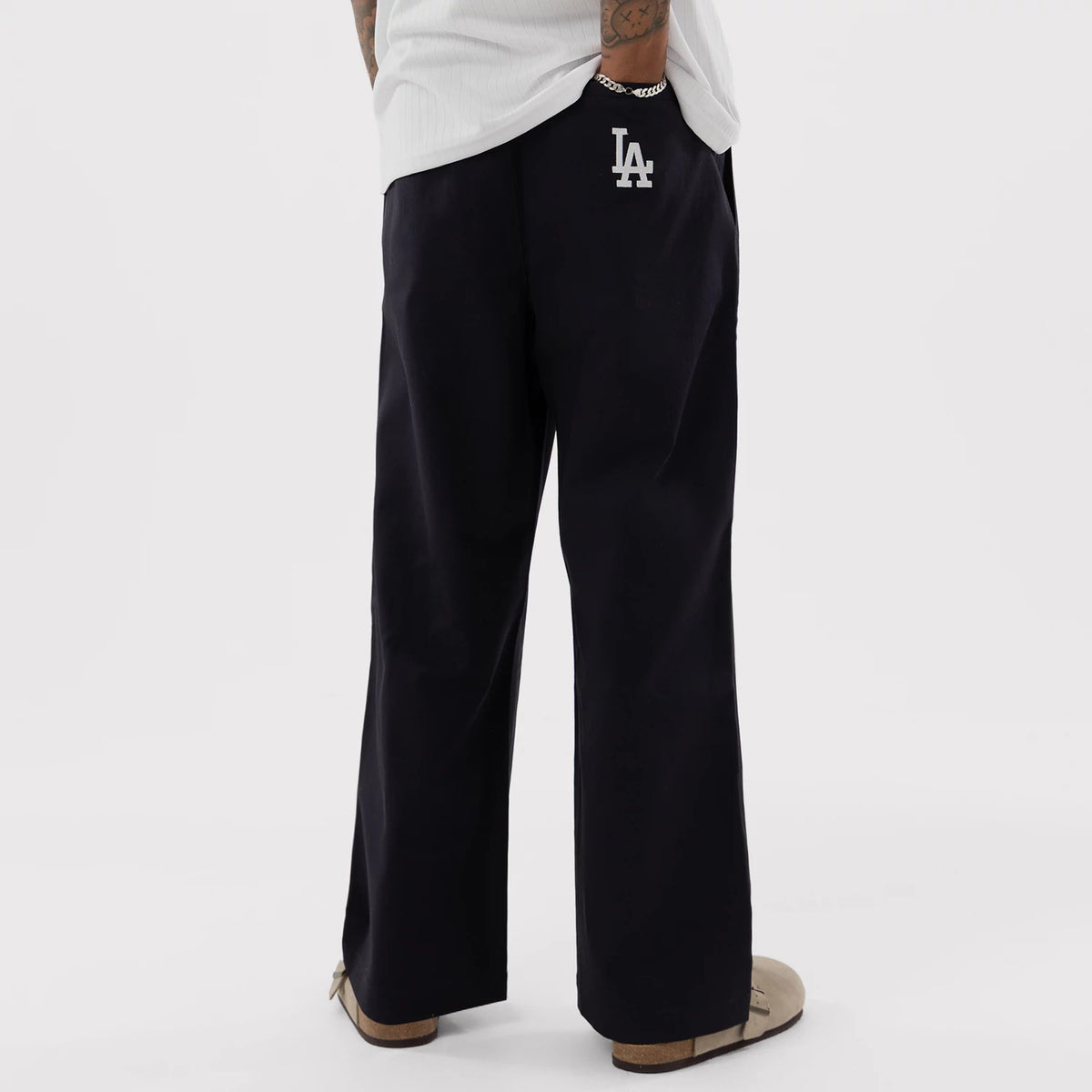 New Era LA Dodgers MLB Navy Woven Chinos, back view on a model showing the embroidered white LA logo on the right pocket, straight-leg fit with a loose silhouette, and a casual styling with beige slip-on shoes and a simple white shirt.