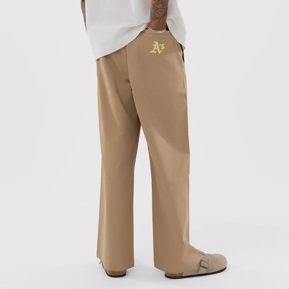 New Era Athletics MLB Beige Woven Chinos, back view on a model showing the embroidered yellow A's logo on the right pocket, straight leg fit, and the beige fabric with a slightly flared hem worn with casual shoes.