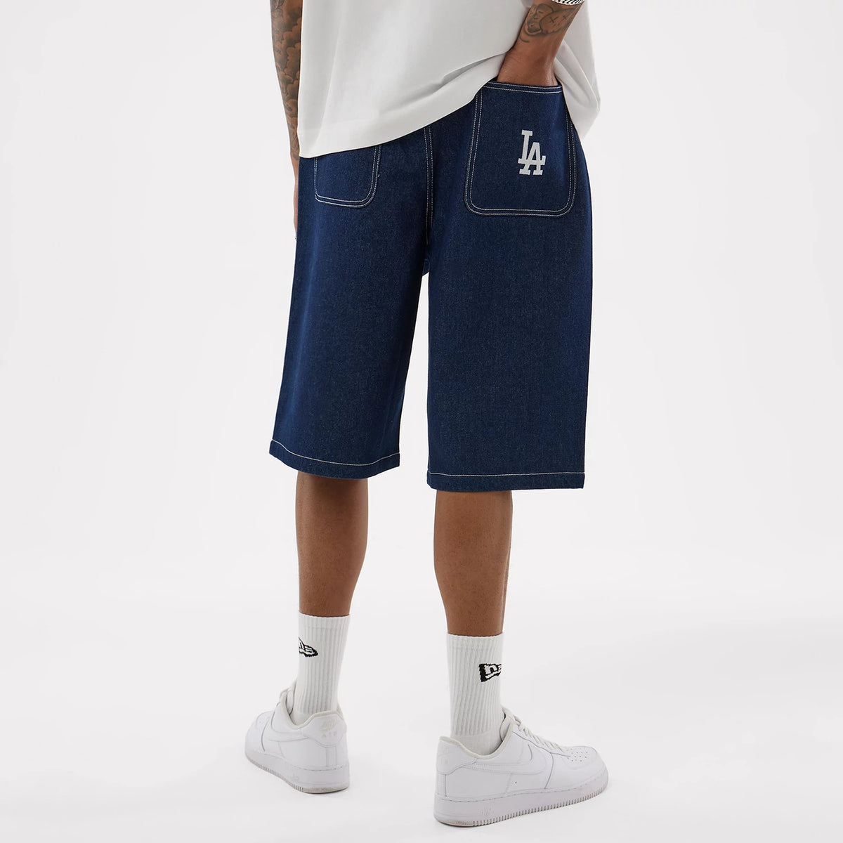 New Era LA Dodgers MLB Jort Dark Blue Oversized Shorts, back view showing the white embroidered LA Dodgers logo on the right pocket, top stitching across both back pockets, styled casually with white trainers and socks featuring the New Era logo.