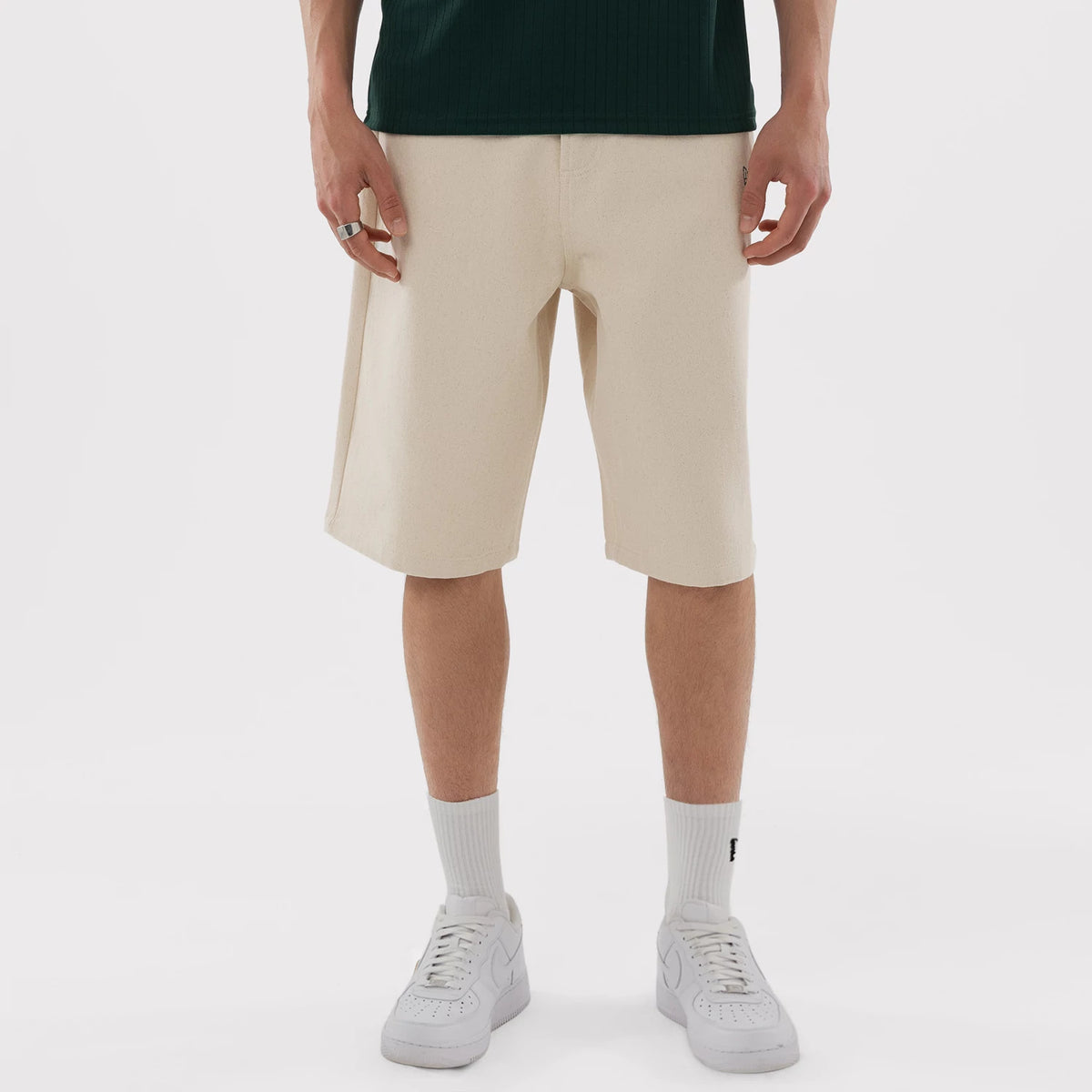 New Era Athletics MLB Jort Cream Oversized Shorts, front view on-model showing the cream-coloured knee-length design with a straight cut silhouette, a embroidered logo on the hem and styled with a dark top and white trainers.