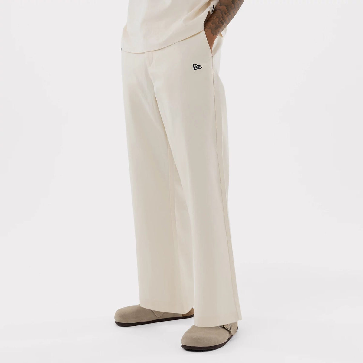 New Era New York Yankees MLB Cream Woven Chinos, on-model view showcasing a relaxed fit with straight legs, featuring an embroidered New Era flag logo on the left pocket and styled with taupe suede shoes and a coordinating cream top.