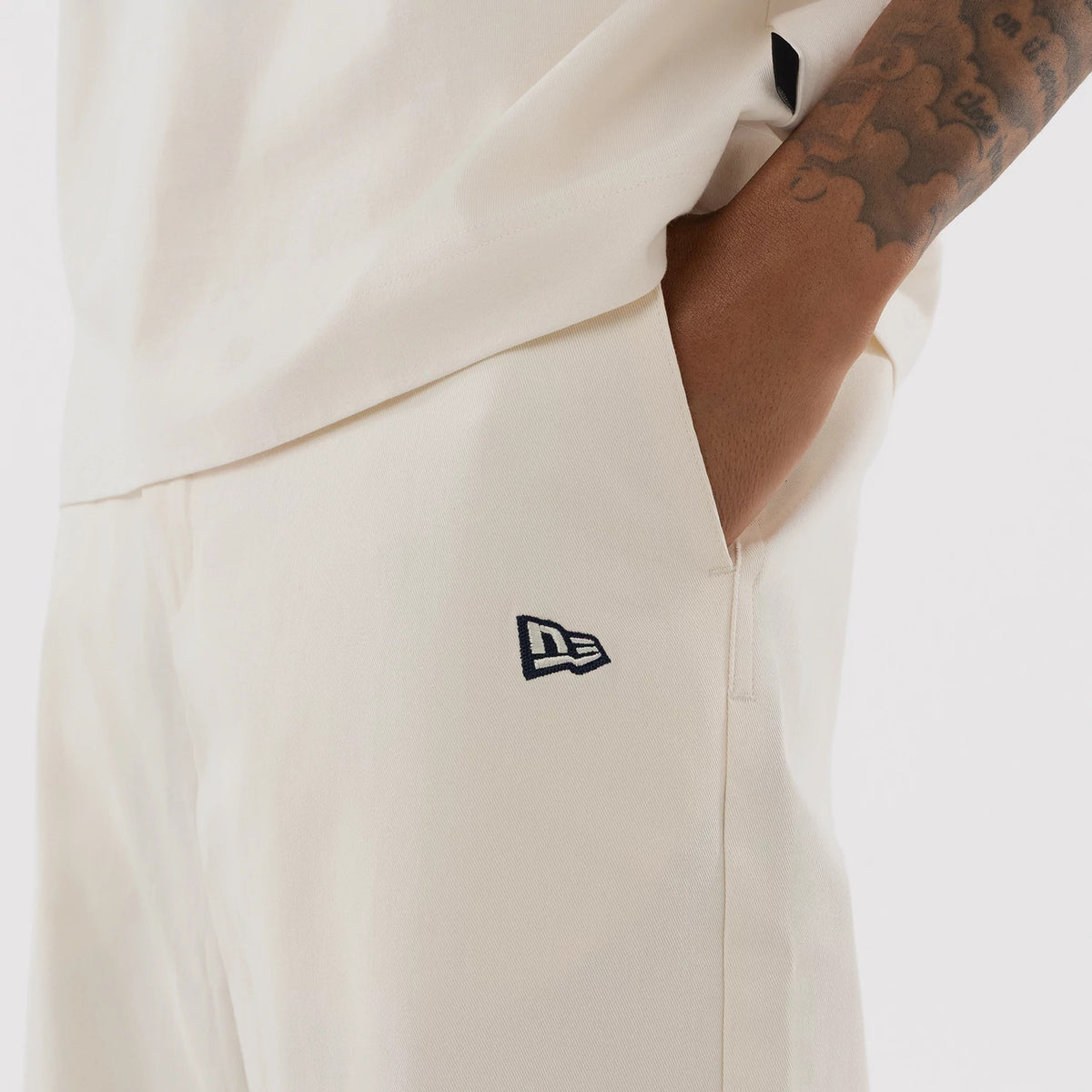 New Era New York Yankees MLB Cream Woven Chinos, side view showing the navy embroidered New Era flag logo above the pocket, a cream fabric with a texture, and a close-up of a hand resting in the pocket against a short-sleeved top.
