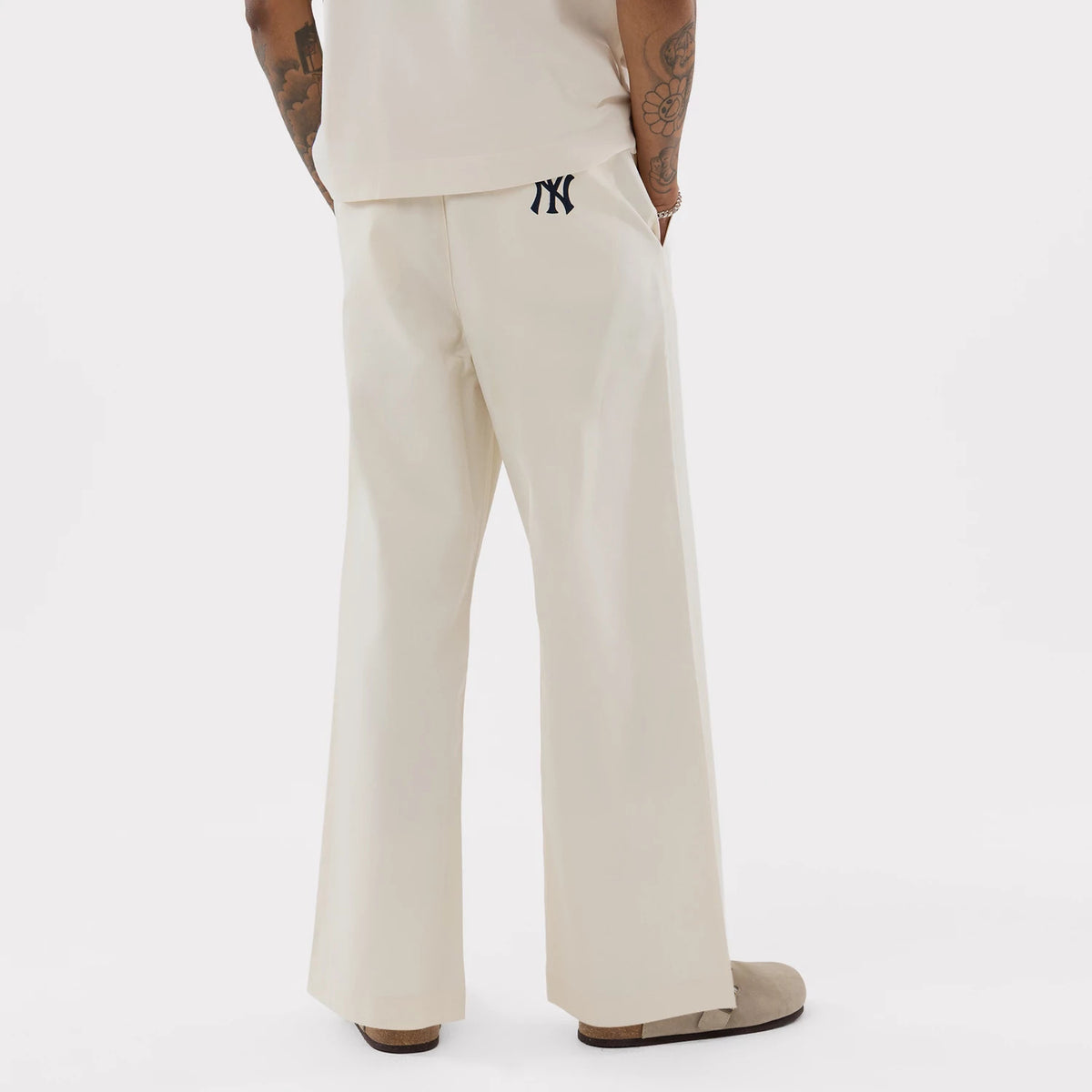 New Era New York Yankees MLB Cream Woven Chinos, rear view on a model showing the black embroidered NY logo below the waistband, loose-fitting legs, and wide hem falling over light brown shoes.