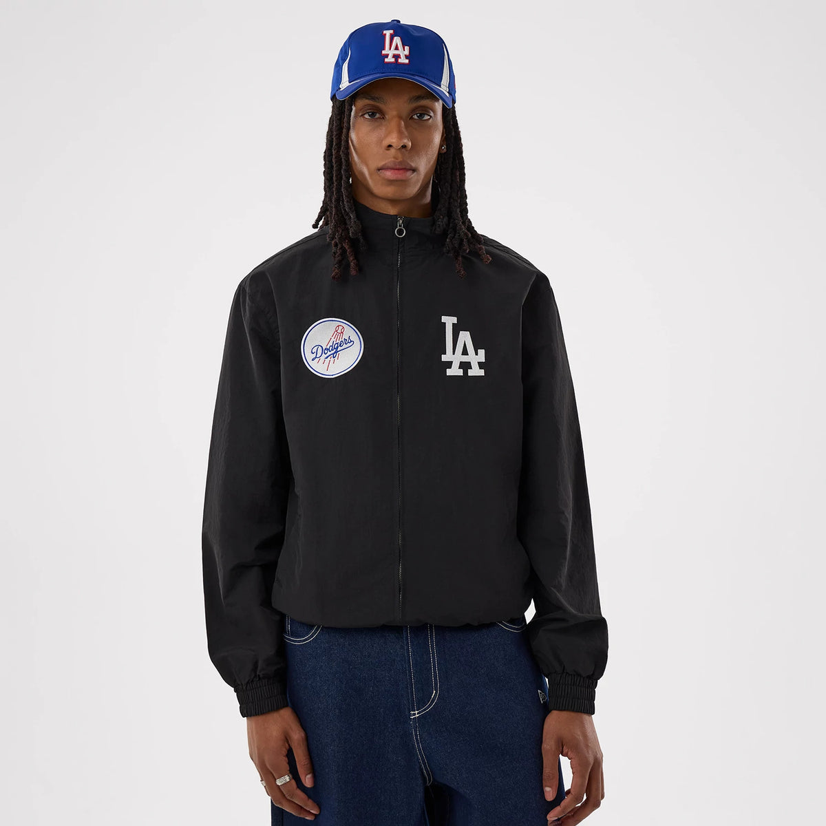 New Era LA Dodgers MLB Black Sports Jacket, front view on a model wearing a blue cap, showing a large white embroidered LA logo on the chest, an oval Dodgers patch on the opposite side, and a full-length zip closure down the centre.
