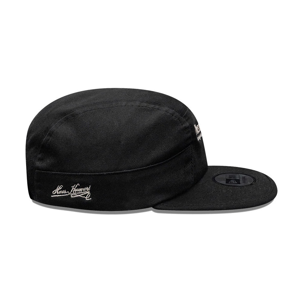 New Era X Hermanos Koumori Running Black Camper Cap, side view showing the white Hermanos Koumori script logo embroidered on the side panel, a flat black visor with a round sticker on top, and a curved black crown.