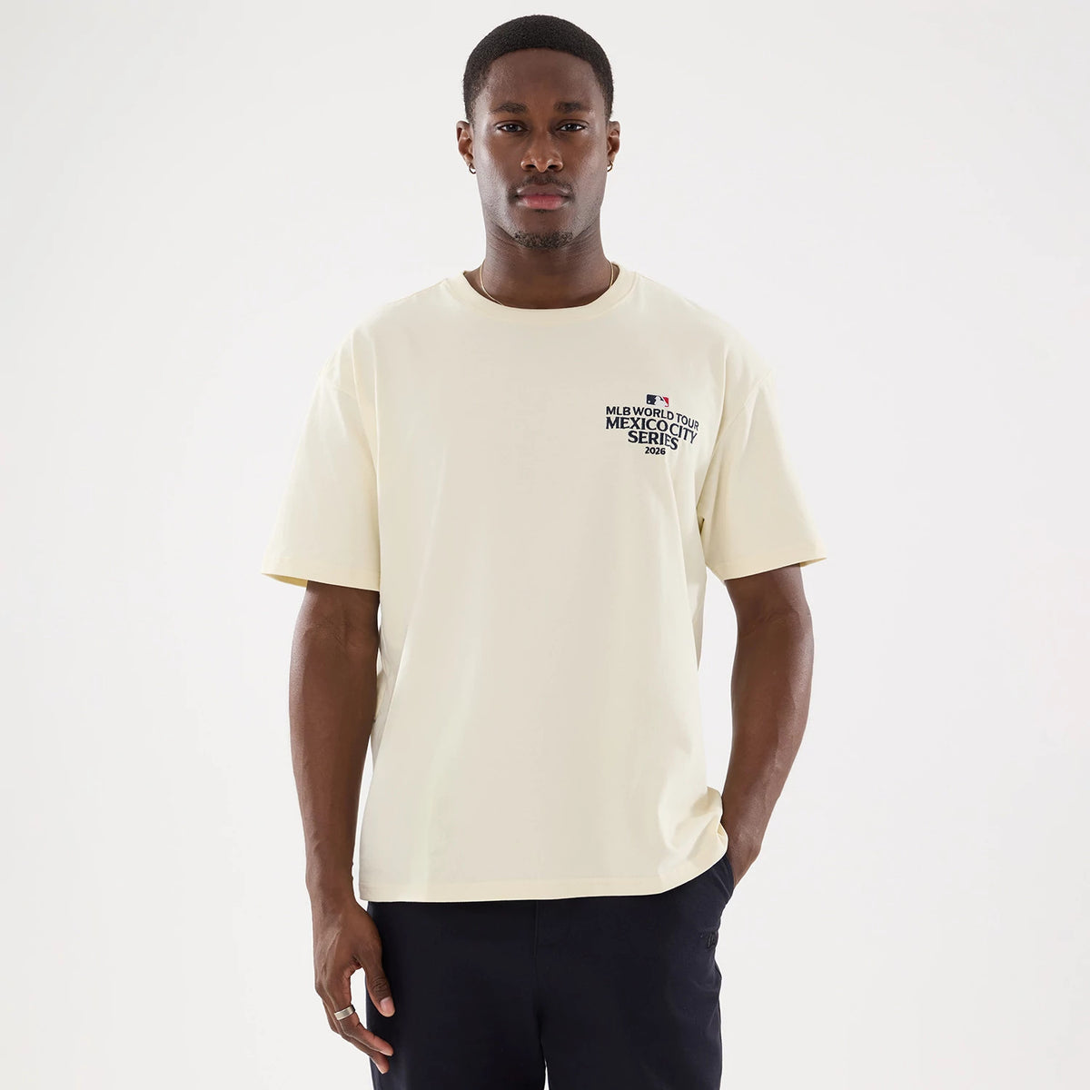 New Era Dueling Logo MLB Mexico City Series 2026 Script Cream T-Shirt, front view on a model showing the cream-coloured short-sleeved tee with a small printed MLB World Tour Mexico City Series 2026 logo on the left chest, styled with dark trousers.