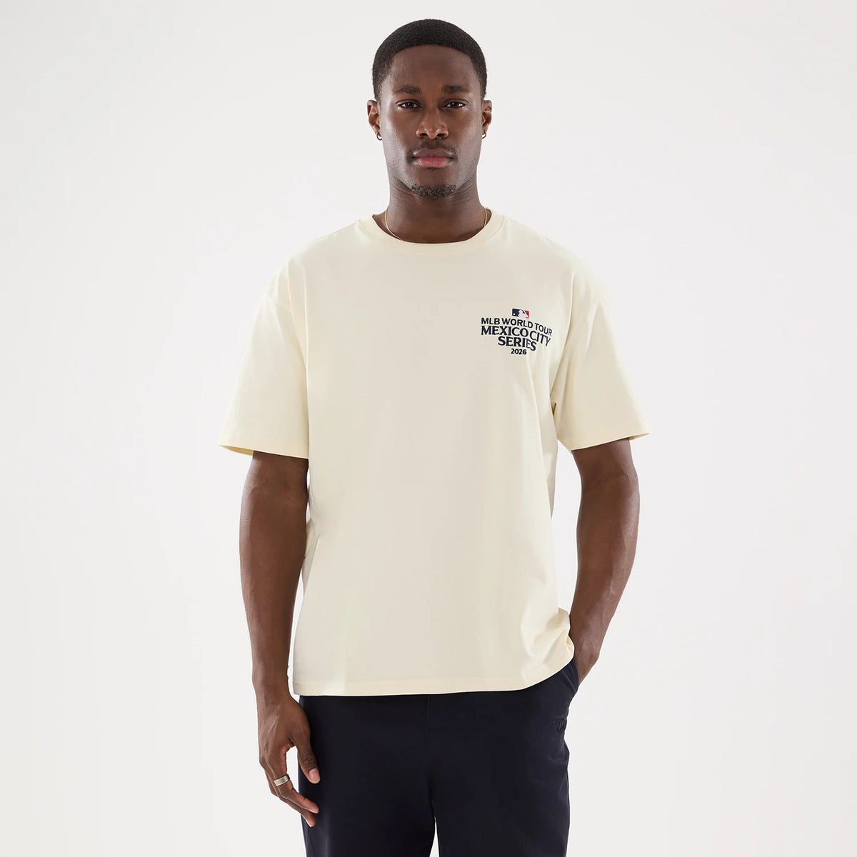 New Era Dueling Logo MLB Mexico City Series 2026 Script Cream T-Shirt, front view on a model showcasing a cream-coloured tee with a printed black and red logo on the left chest, styled with dark trousers for a casual look.