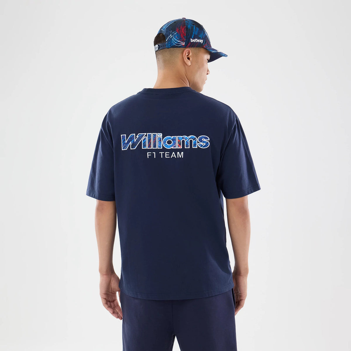 New Era Williams GB Race Special Navy T-Shirt, back view on a model with a coordinating cap, displaying large Williams F1 Team lettering across the upper back in a patterned design, set against the plain navy fabric of the t-shirt.