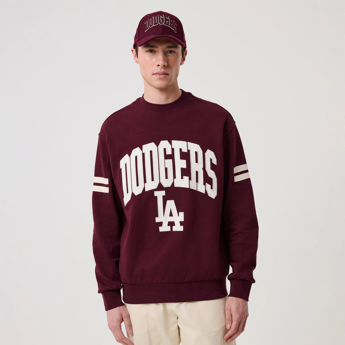 New Era LA Dodgers MLB Arch Graphic Dark Purple Crew Neck Sweater, front view on a model with a matching cap, featuring white Dodgers lettering across the chest, white LA logo below, and two white stripes on each sleeve for detail.